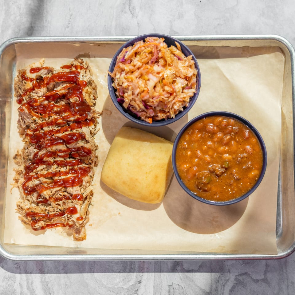 Overhead view of barbecue platter with sauced pulled pork, coleslaw, baked beans, and two golden cornbread pieces on a metal tray.