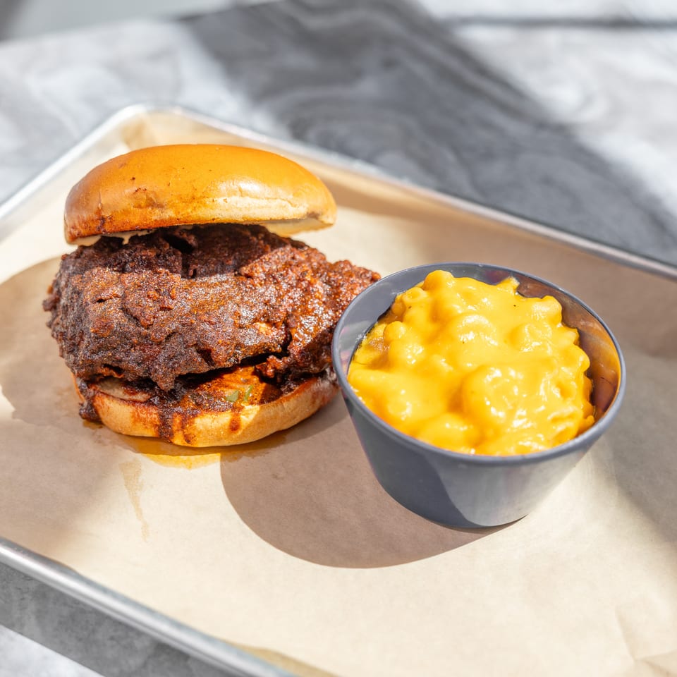 A crispy fried chicken sandwich on a brioche bun sits beside a bowl of creamy macaroni and cheese on a lined tray.