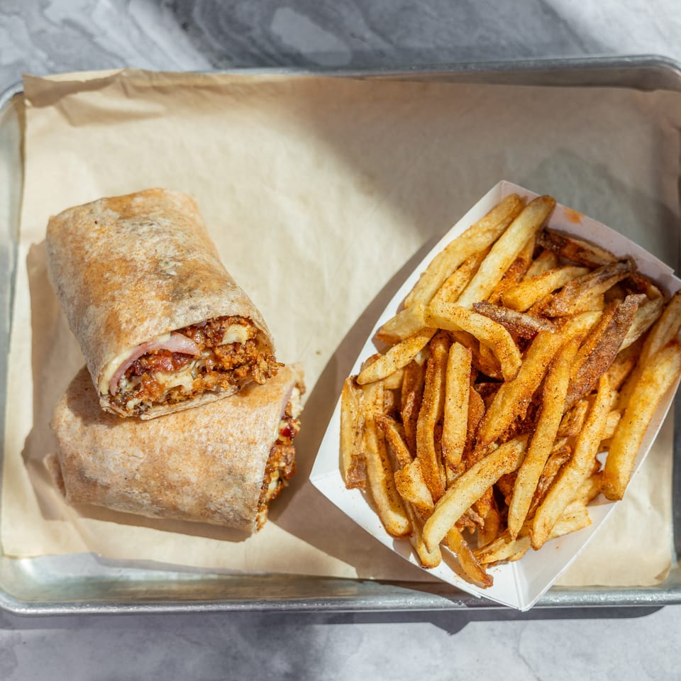 A toasted wrap filled with meat, cheese, and vegetables sits beside seasoned French fries on a parchment-lined metal tray.