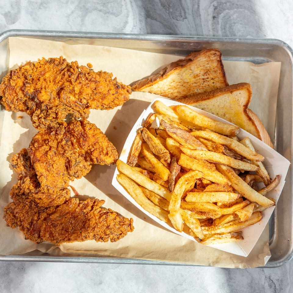 Tray of crispy fried chicken tenders, seasoned fries, and toasted bread served on parchment paper.