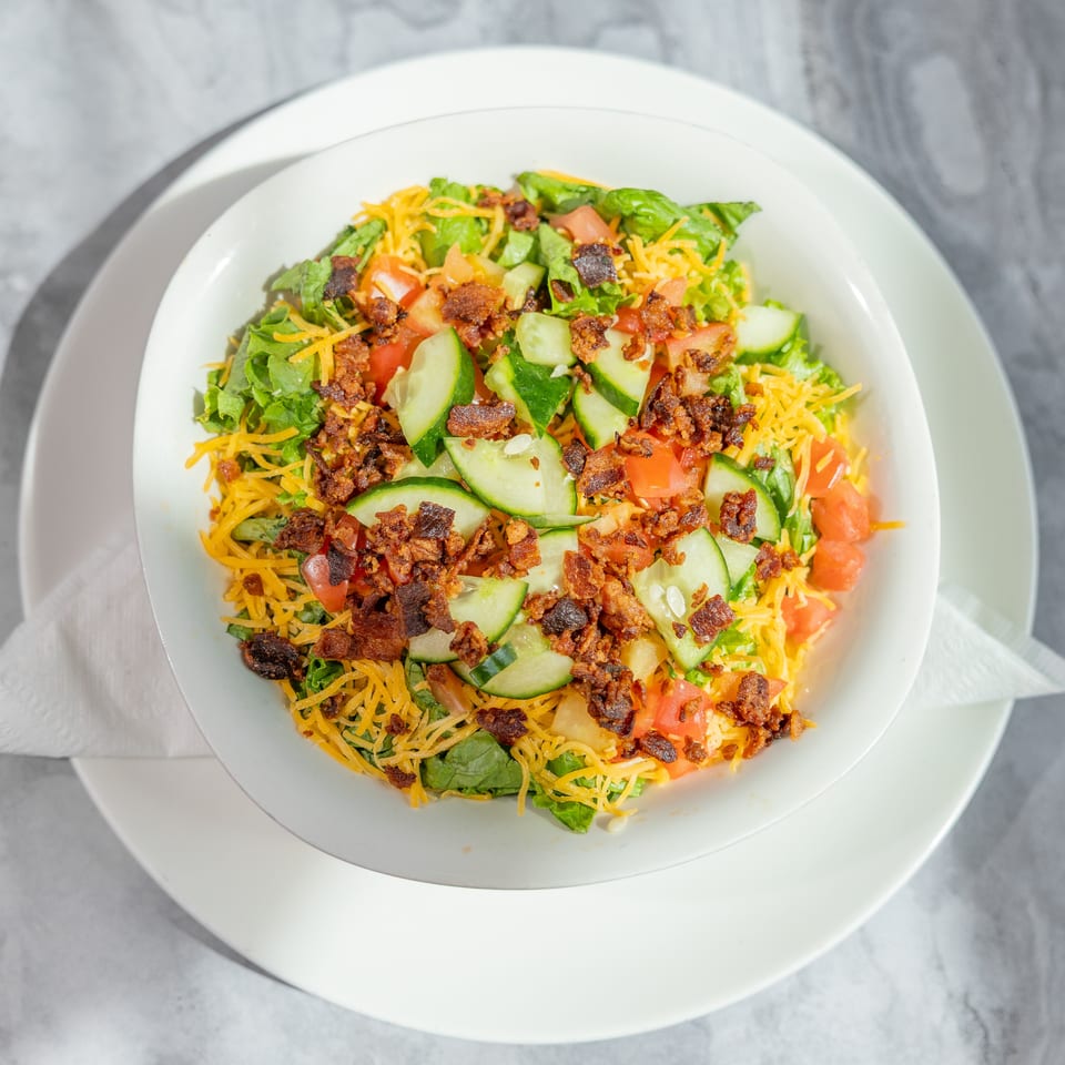 Overhead view of a fresh salad with lettuce, cucumbers, tomatoes, shredded cheddar cheese, and crispy bacon on a white plate.