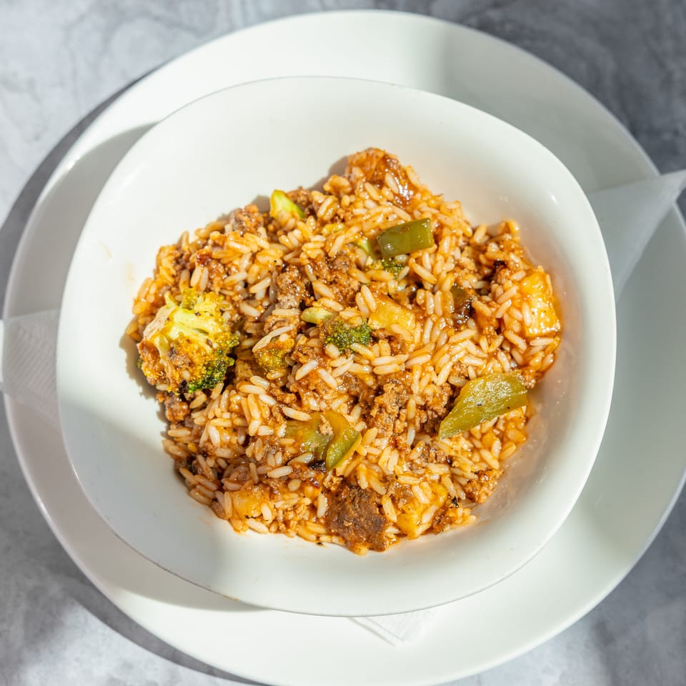 Bowl of saucy rice mixed with ground meat, shrimp, broccoli, and green peppers on a white plate.