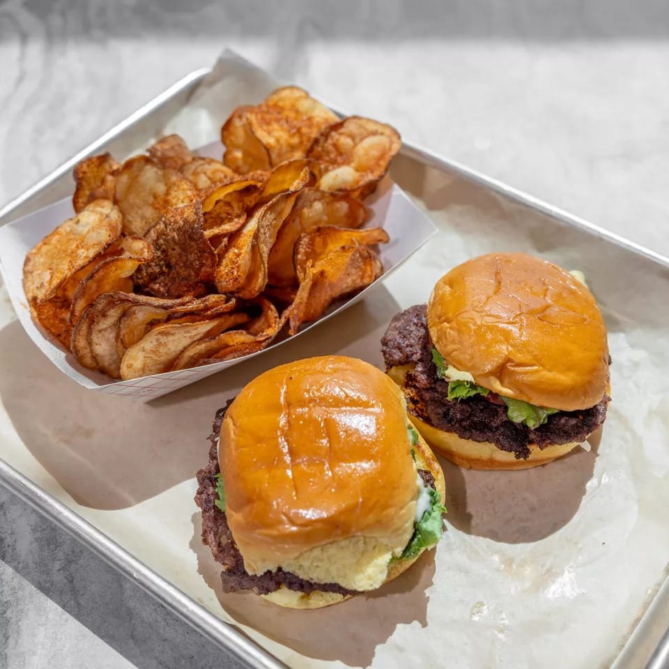 Two cheeseburgers with lettuce and crispy potato chips served on a paper-lined metal tray.