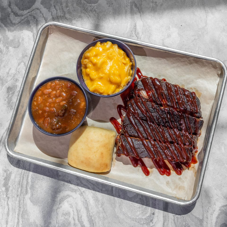 Barbecue ribs drizzled with sauce served with baked beans, macaroni and cheese, and a bread roll on a tray.